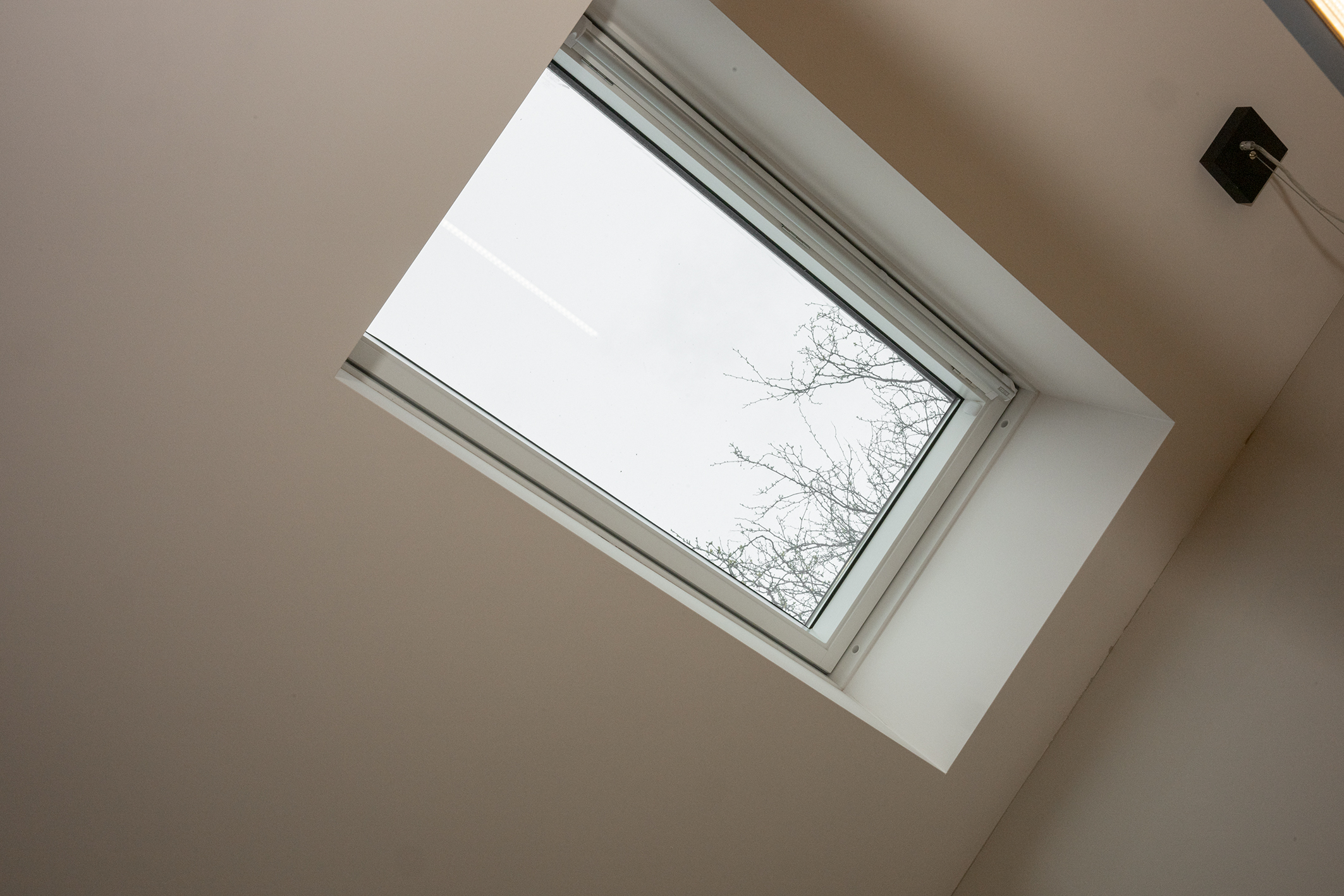 Onderaanzicht van een modern rechthoekig dakraam dat uitkijkt op een bewolkte lucht en boomtakken.