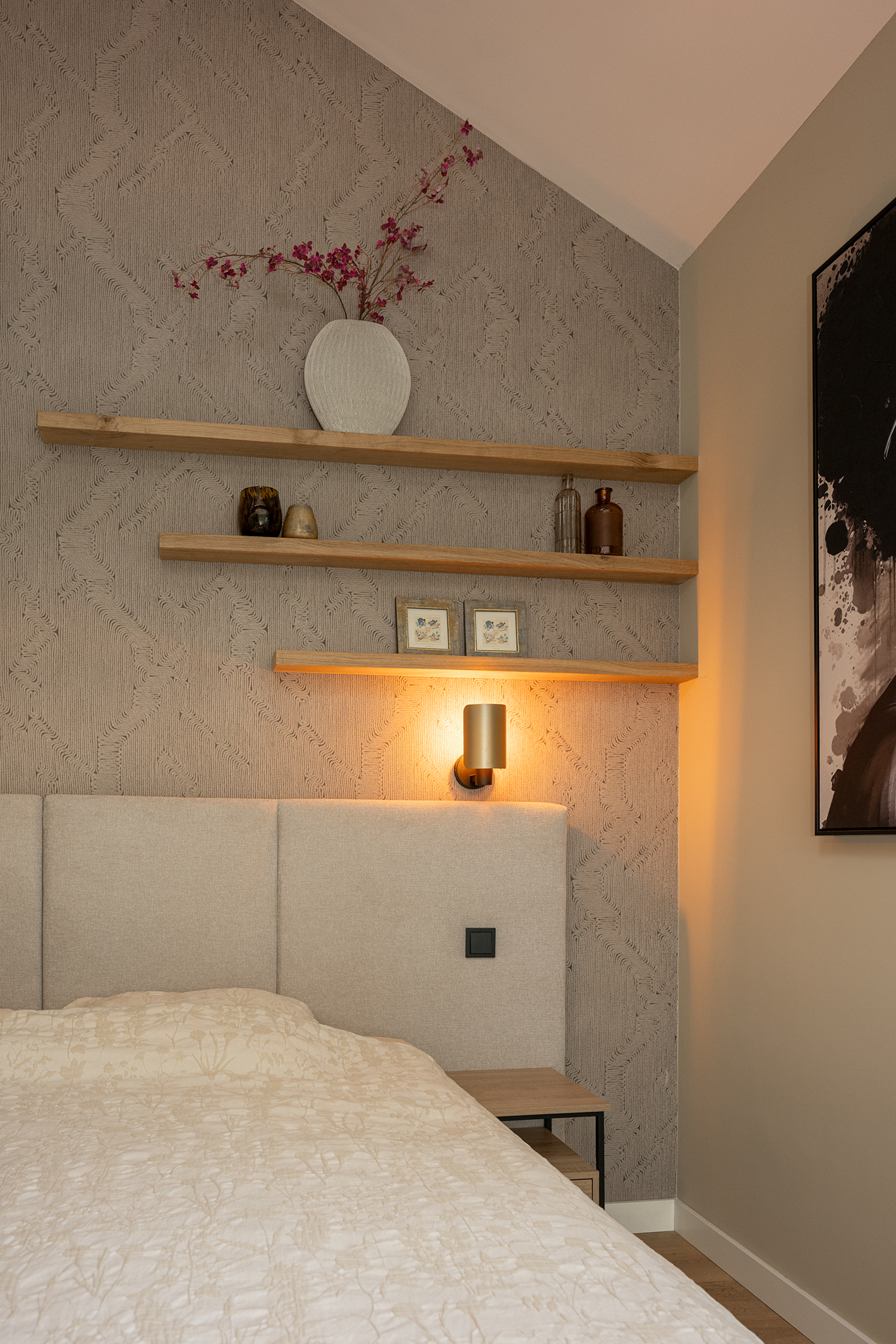 Zwevende houten wandplanken boven een bed in een slaapkamer met getextureerd behang. Een wandlamp verlicht de planken. De vloer is van hout.