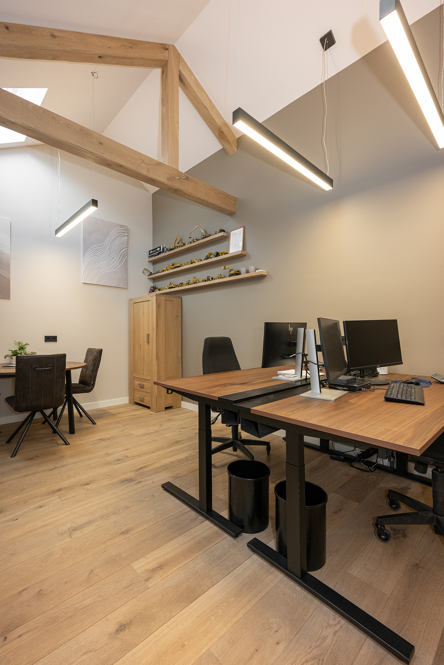 Zijaanzicht van de werkplek met focus op het houten bureau, zwarte bureaustoelen en de rustieke houten steunbalken aan het plafond.