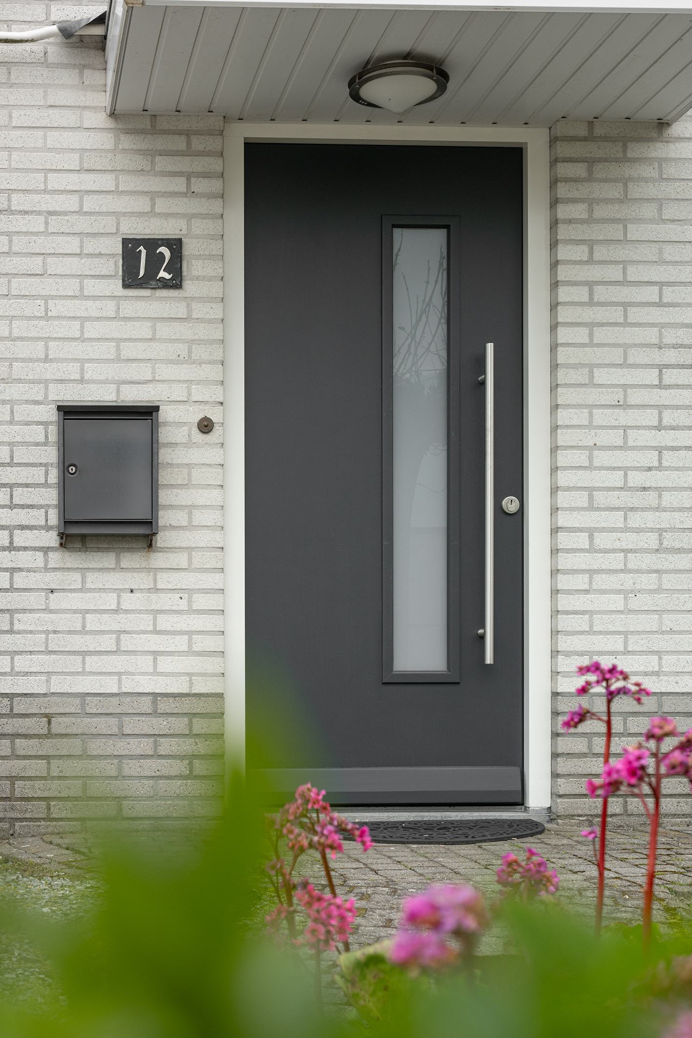 Vooraanzicht van een grijze voordeur met glas, huisnummer 12 en een zwarte wandbrievenbus aan een witte muur.