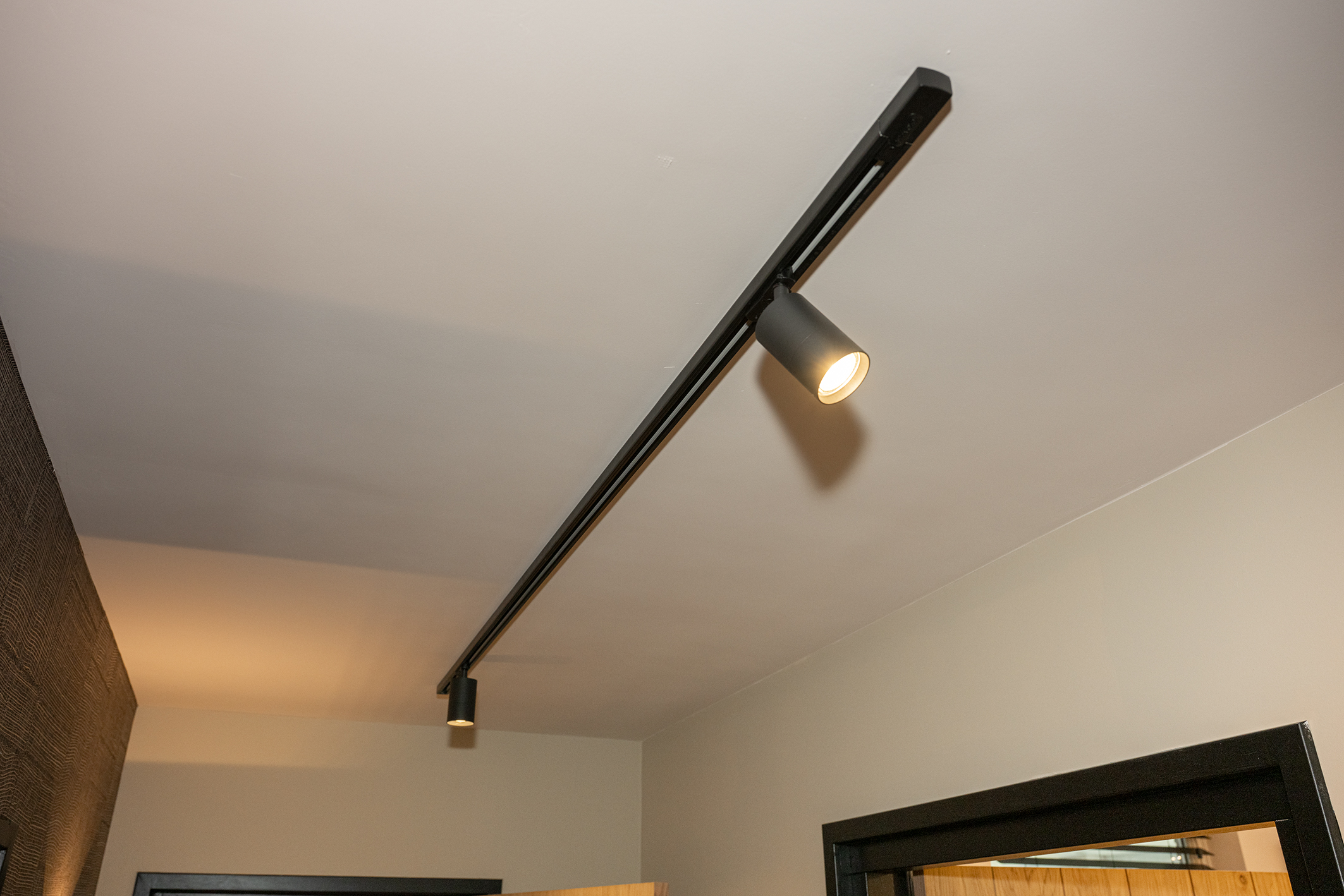 Minimalistische zwarte railverlichting met twee verstelbare spots aan een wit plafond in een gang.