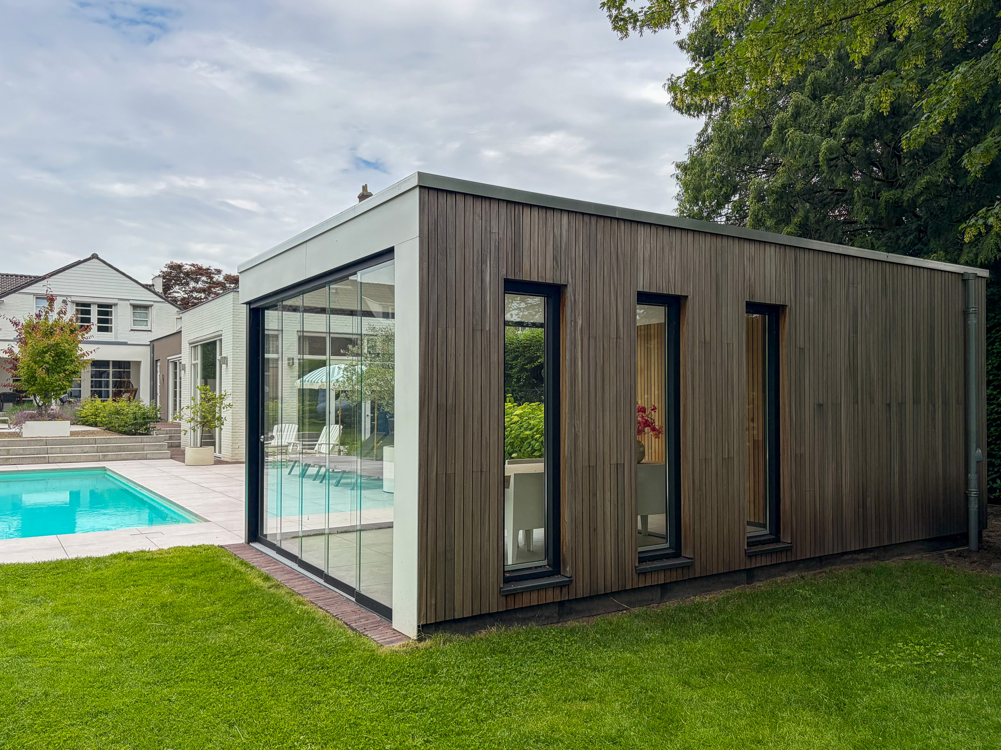 Schuine hoekopname van een design tuinhuis met houten gevelbekleding en glazen wanden, gelegen naast een betegeld terras met zwembad.