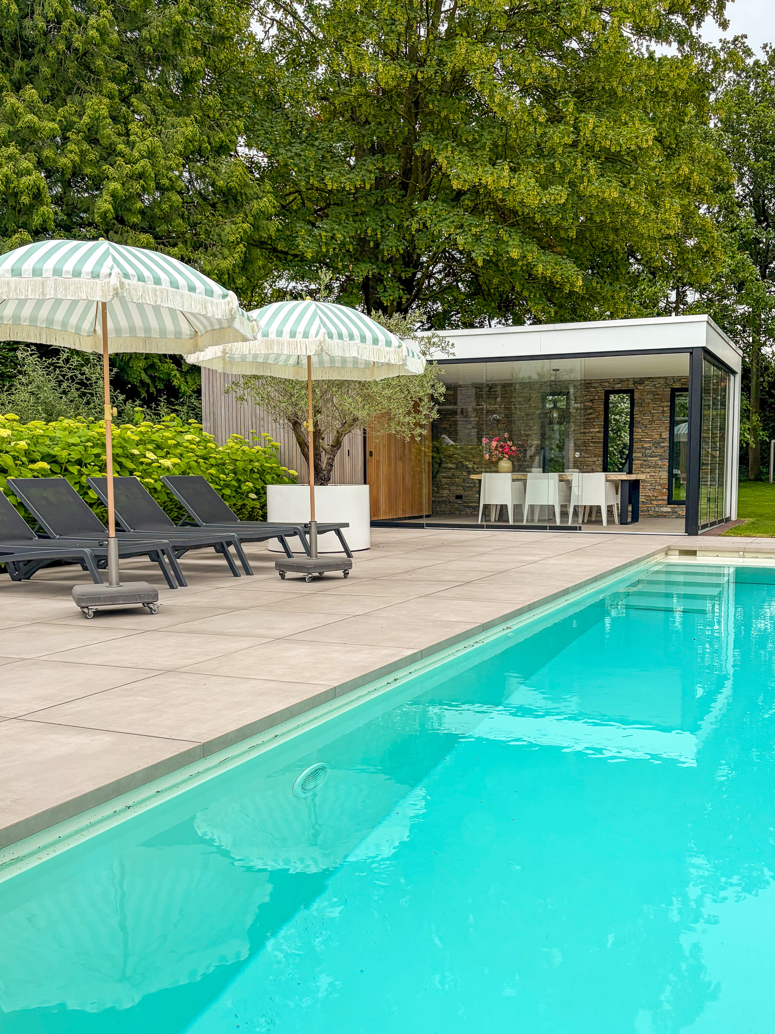 Sfeervolle tuininrichting met een groot zwembad dat het zonlicht reflecteert, geflankeerd door een loungeterras met parasols en een luxe buitenverblijf met natuurstenen wand.