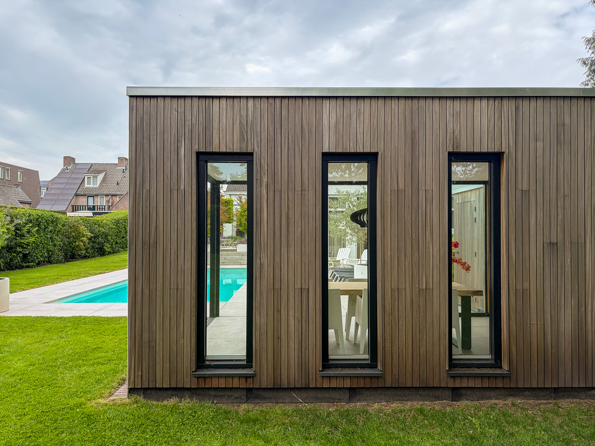 Modern tuinhuis met verticale houten gevelbekleding en drie smalle, hoge ramen die uitzicht bieden op een eettafel binnen en een zwembad in de tuin.