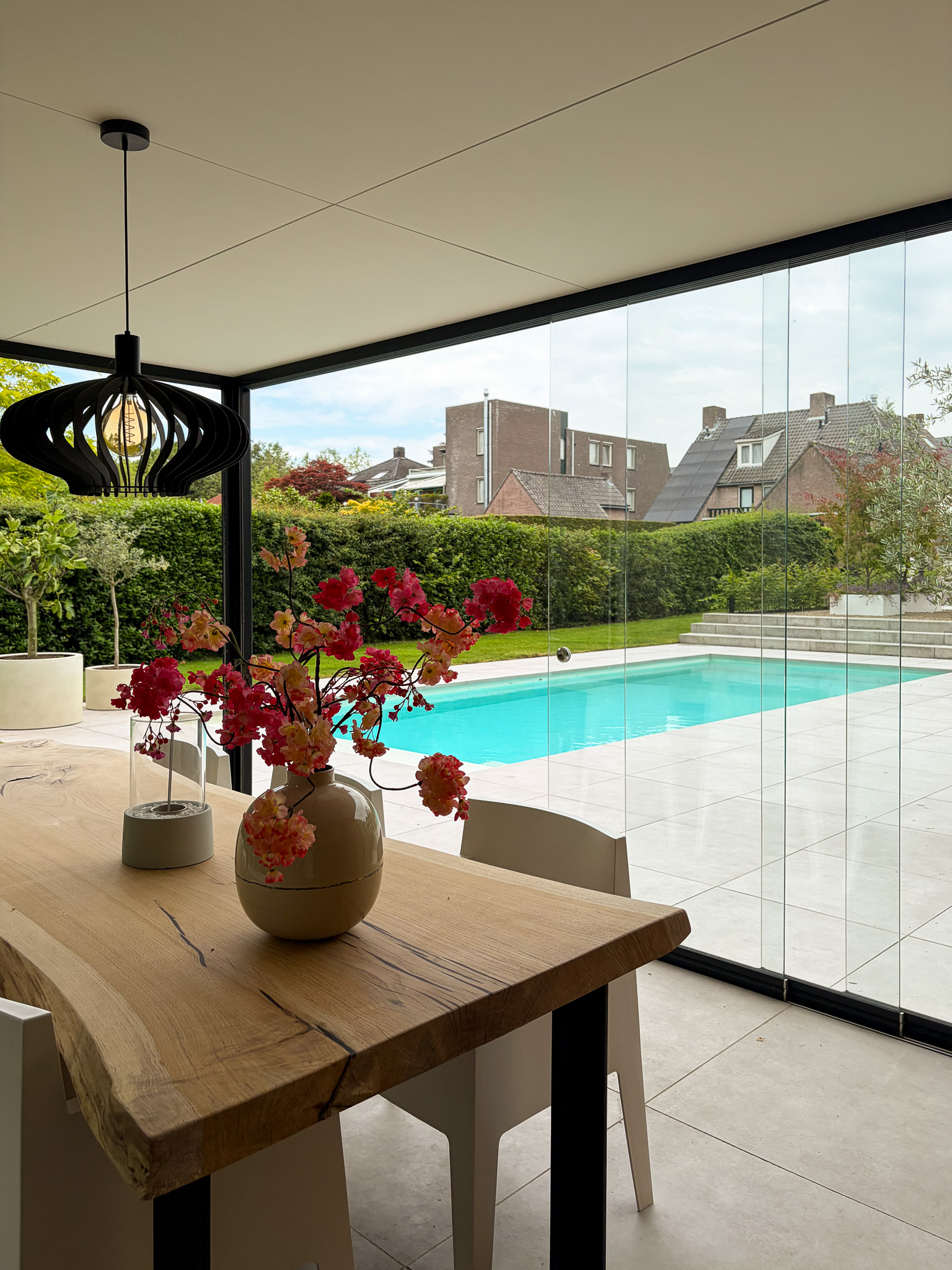 Interieur van een modern tuinkantoor met een robuuste houten eettafel, een vaas met roze kunstbloemen en uitzicht door glas op het zwembad.