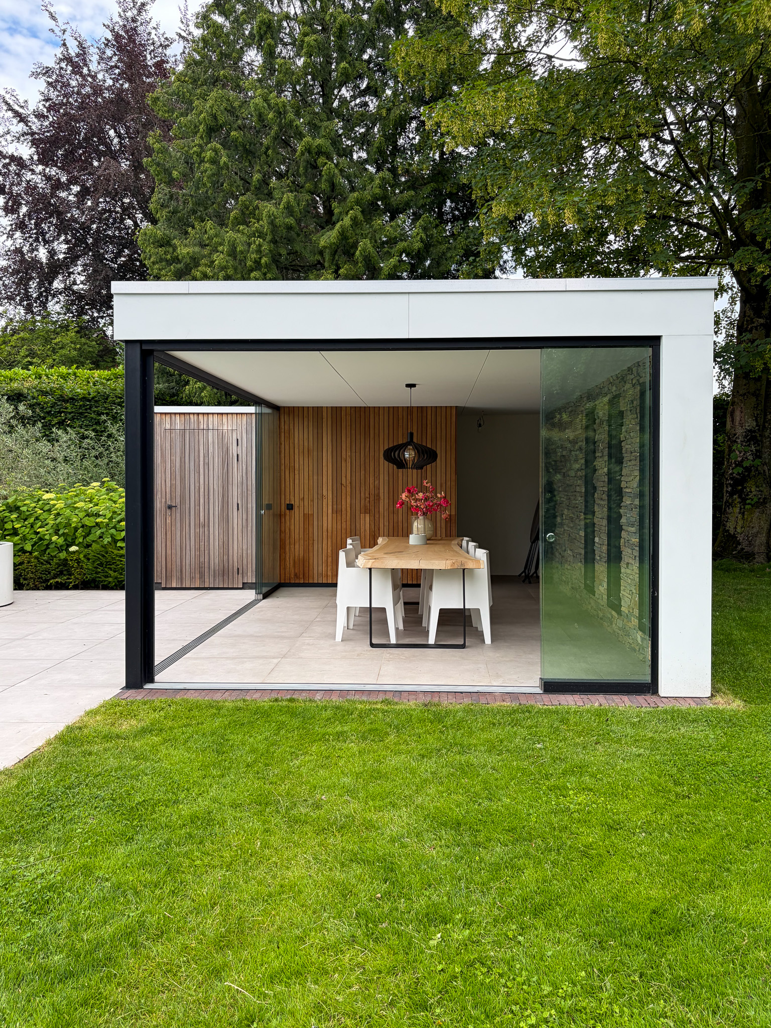 Vooraanzicht van een luxe buitenverblijf met een houten lattenwand, een design hanglamp boven de eethoek en een open glazen schuifsysteem.