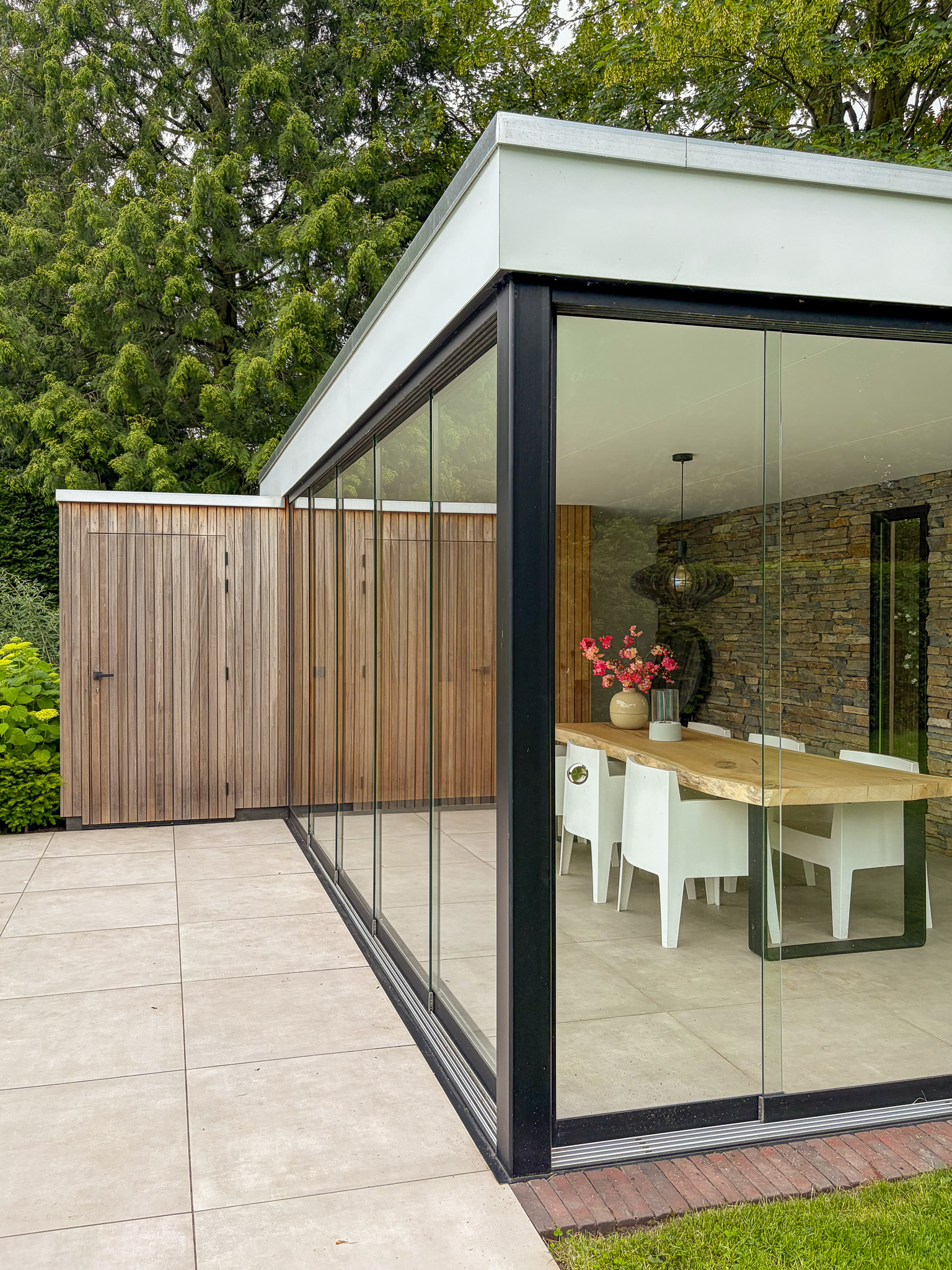 Hoek van een luxe tuinkamer met volledig glazen wanden en een aangrenzende dichte houten berging met een verticale lattenstructuur.