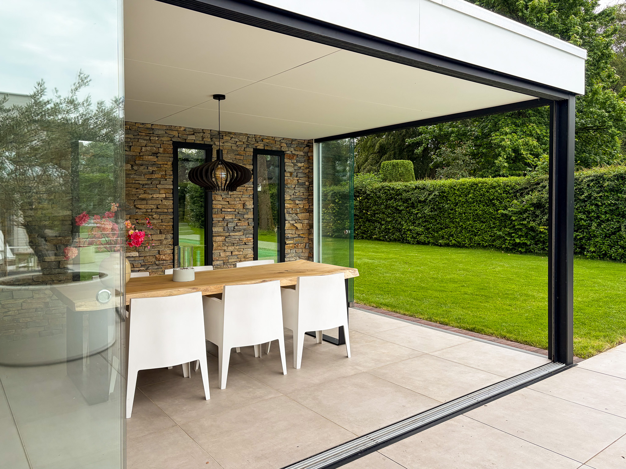 Interieur van een luxe tuinkamer met houten eettafel en witte stoelen, gezien vanaf het terras door een wijd geopende glazen pui.