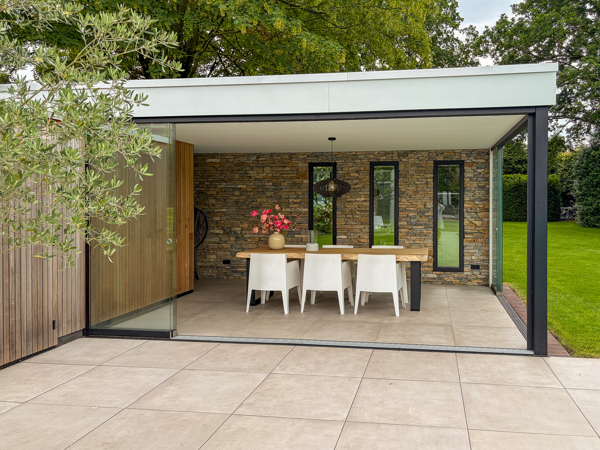 Complete blik op een luxe, open tuinkamer met een strak betegeld terras ervoor, ingericht met een eethoek tegen een robuuste natuurstenen wand onder een wit dak.