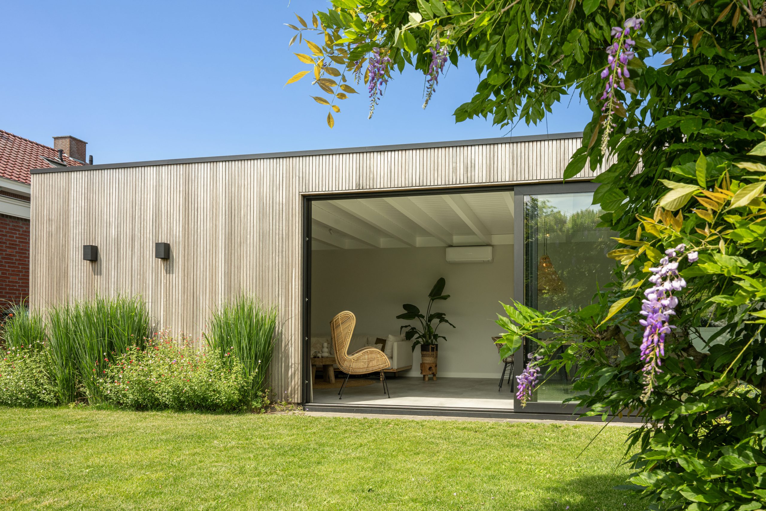 Modern houten tuinkantoor in een groene tuin, sfeervol omringd door bloeiende paarse wisteria (blauweregen) aan de rechterzijde.