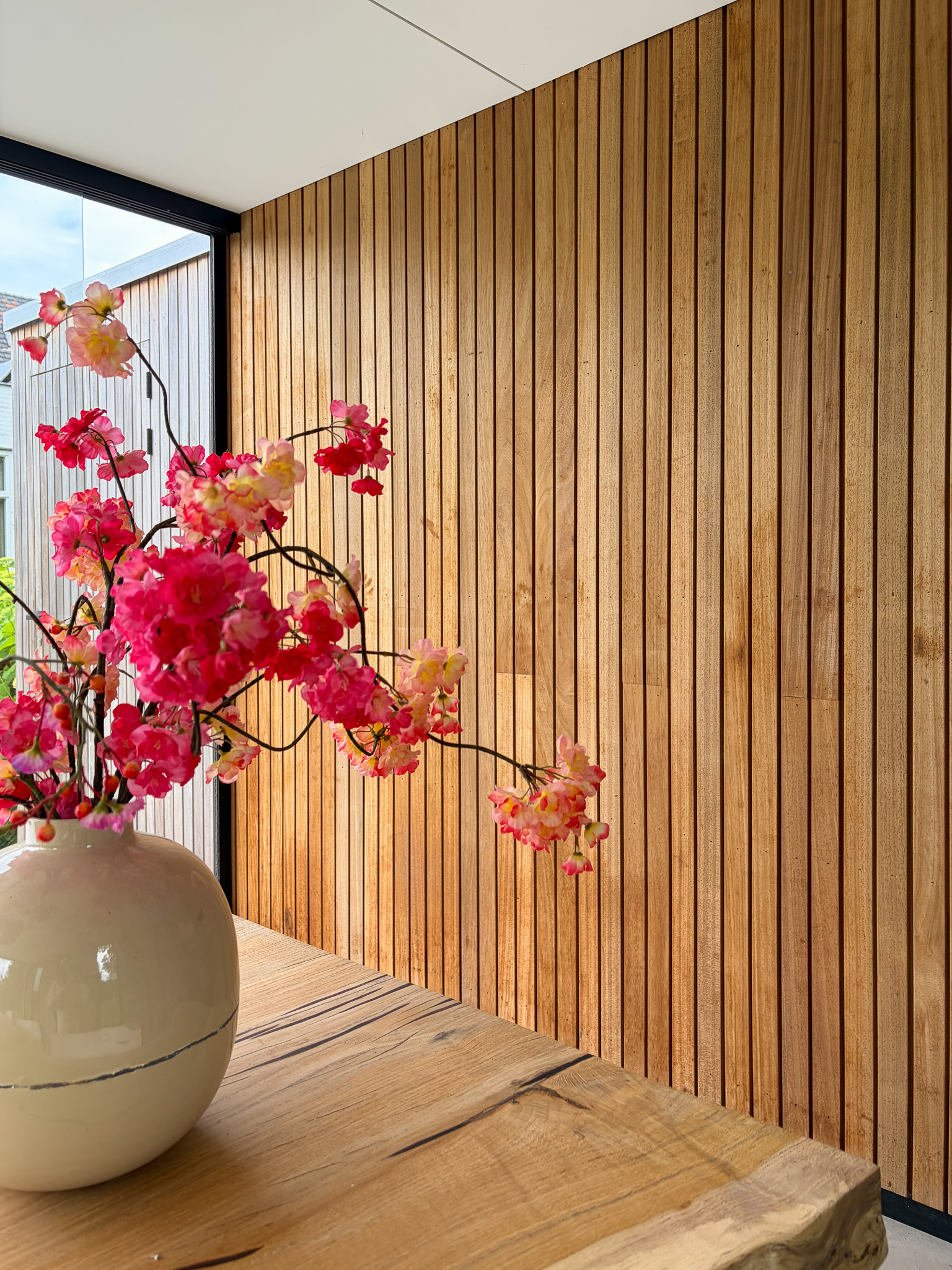 Close-up van een beige vaas met felroze bloesemtakken op een houten tafel tegen een achtergrond van verticale houten latten.