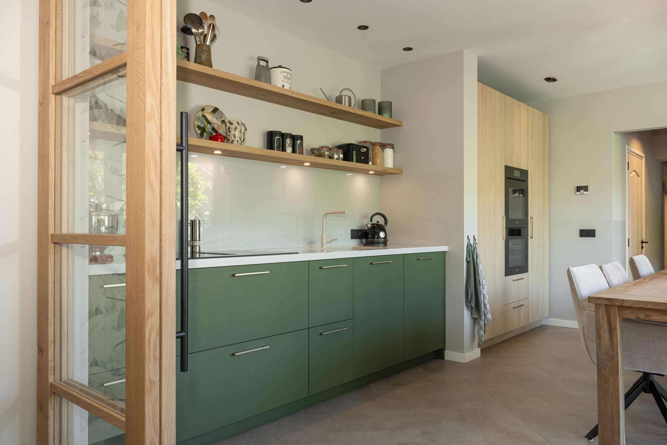 Close-up van een moderne keuken met groene keukenkasten, wit aanrechtblad en open houten wandplanken.