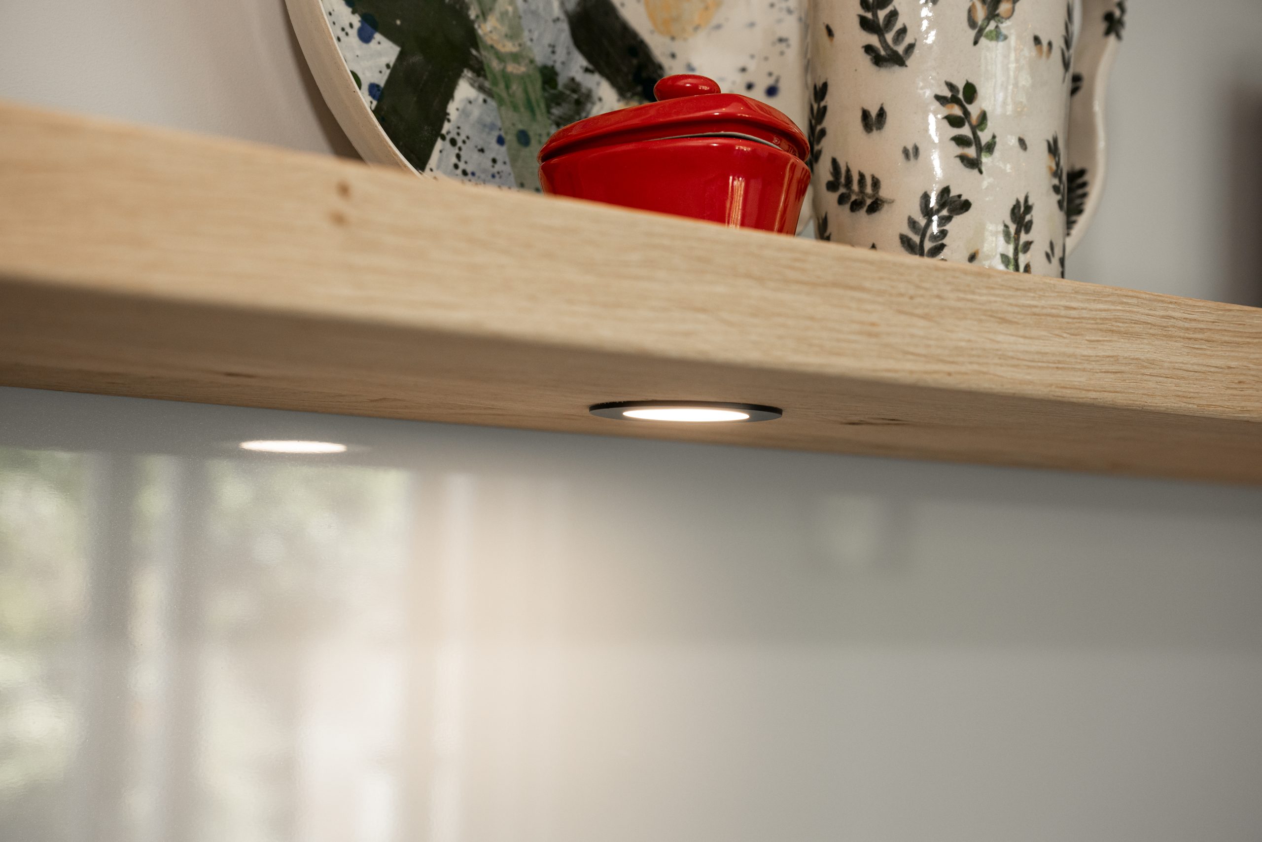 Detailopname van subtiele inbouwverlichting onder een houten legplank in de keuken met decoratieve serviesstukken.