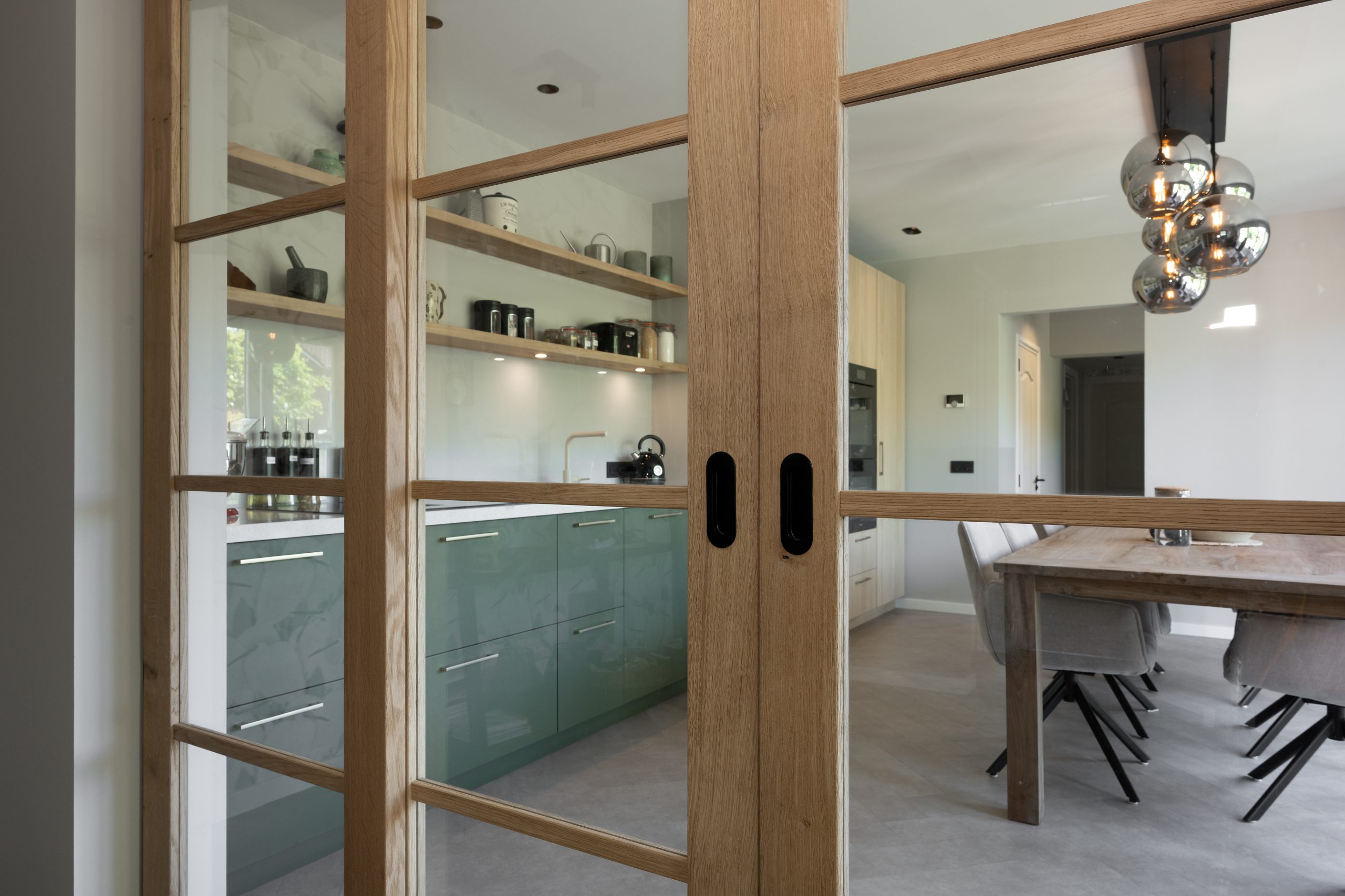 Detailopname van een glazen tussendeur met houten frame, geplaatst naast een moderne groene keuken.
