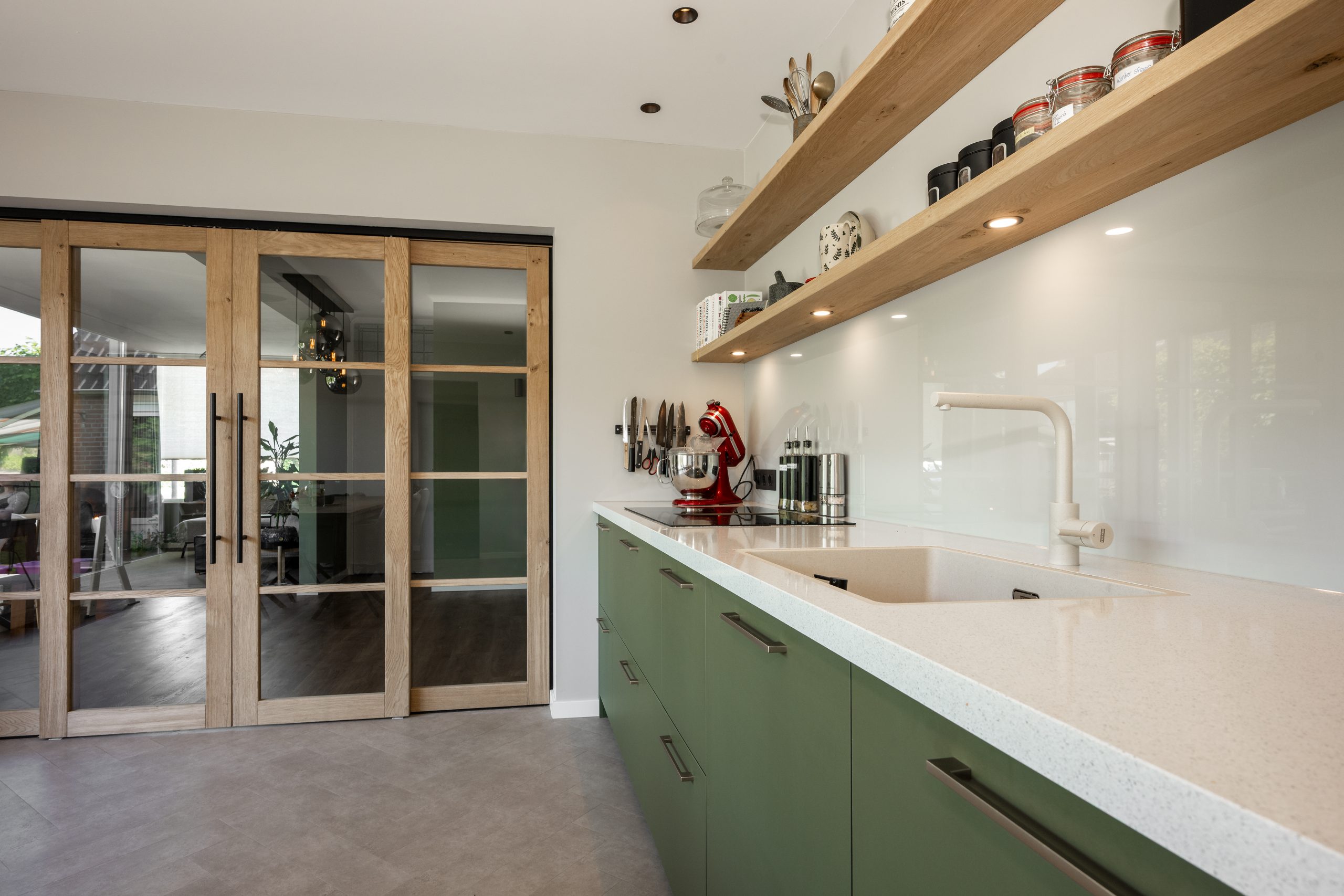 Lichte keukenruimte met groene onderkasten, wit aanrechtblad, houten open planken en decoratieve details.