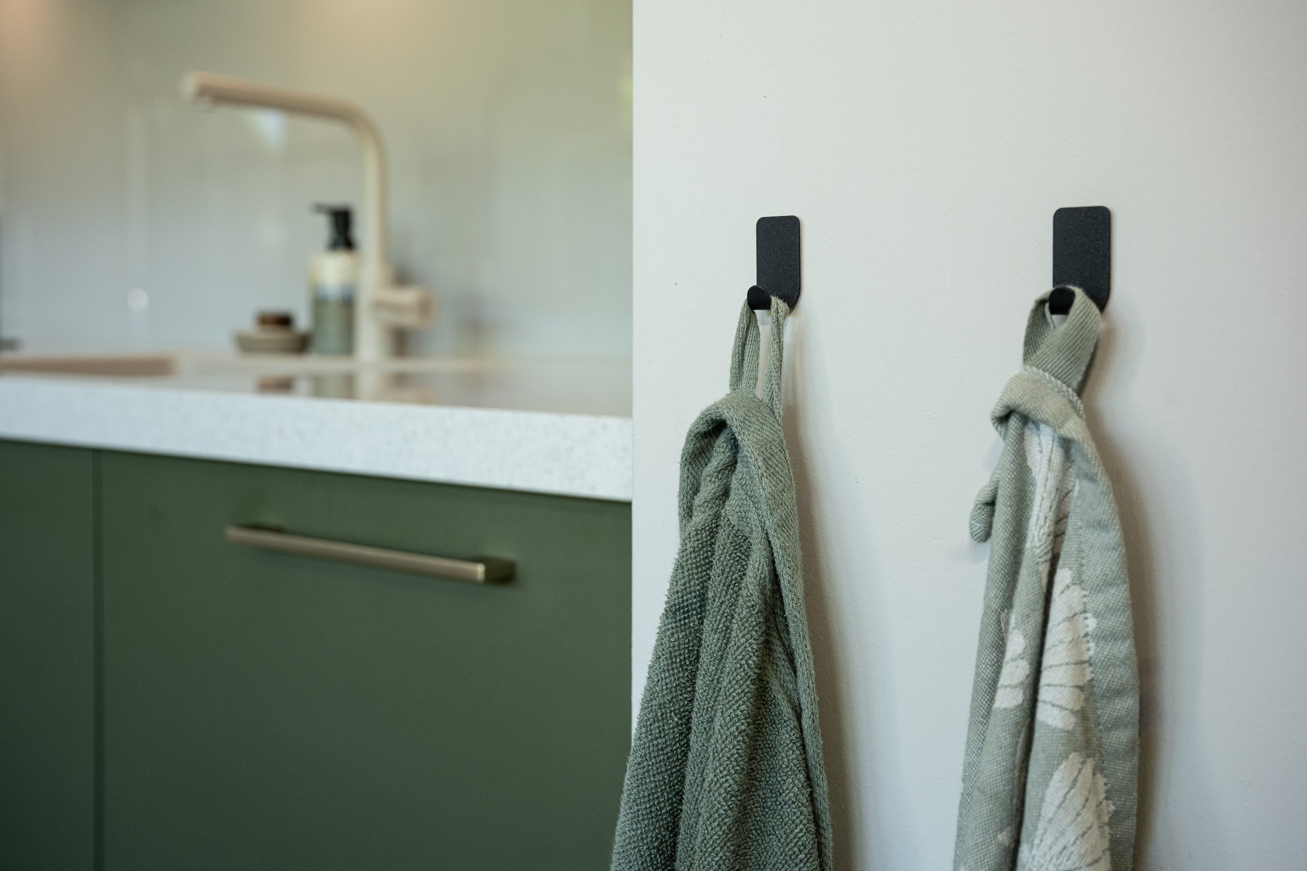 Zwarte wandhaken met keukentextiel, gemonteerd op de muur naast het groene keukenblok.