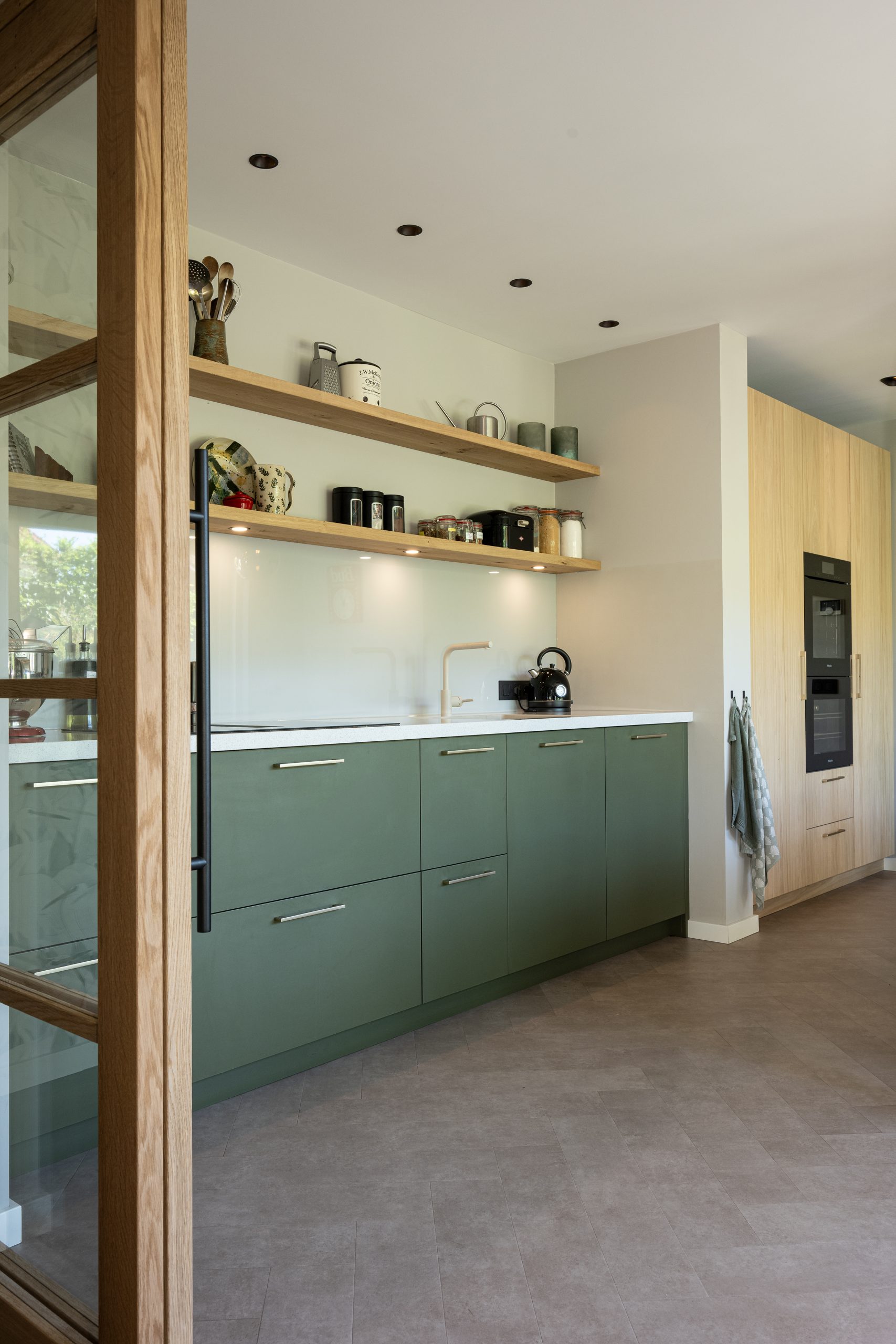 Een moderne groene keukenopstelling in een hoek, gedeeltelijk afgescheiden door een glazen tussendeur met houten frame.