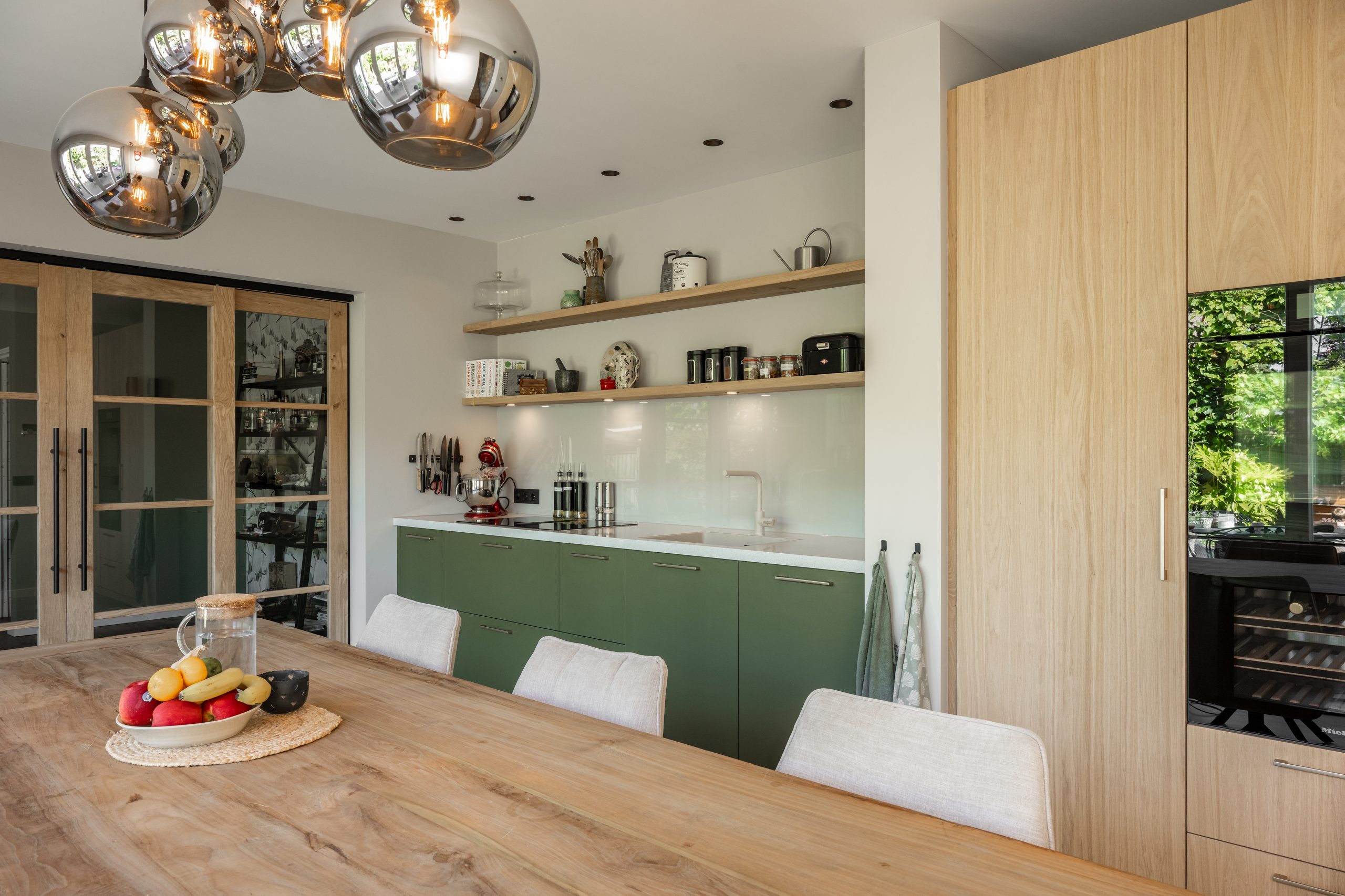 Eethoek met een robuuste houten tafel, luxe pendellampen en uitzicht op een groene keuken met glazen tussendeuren.
