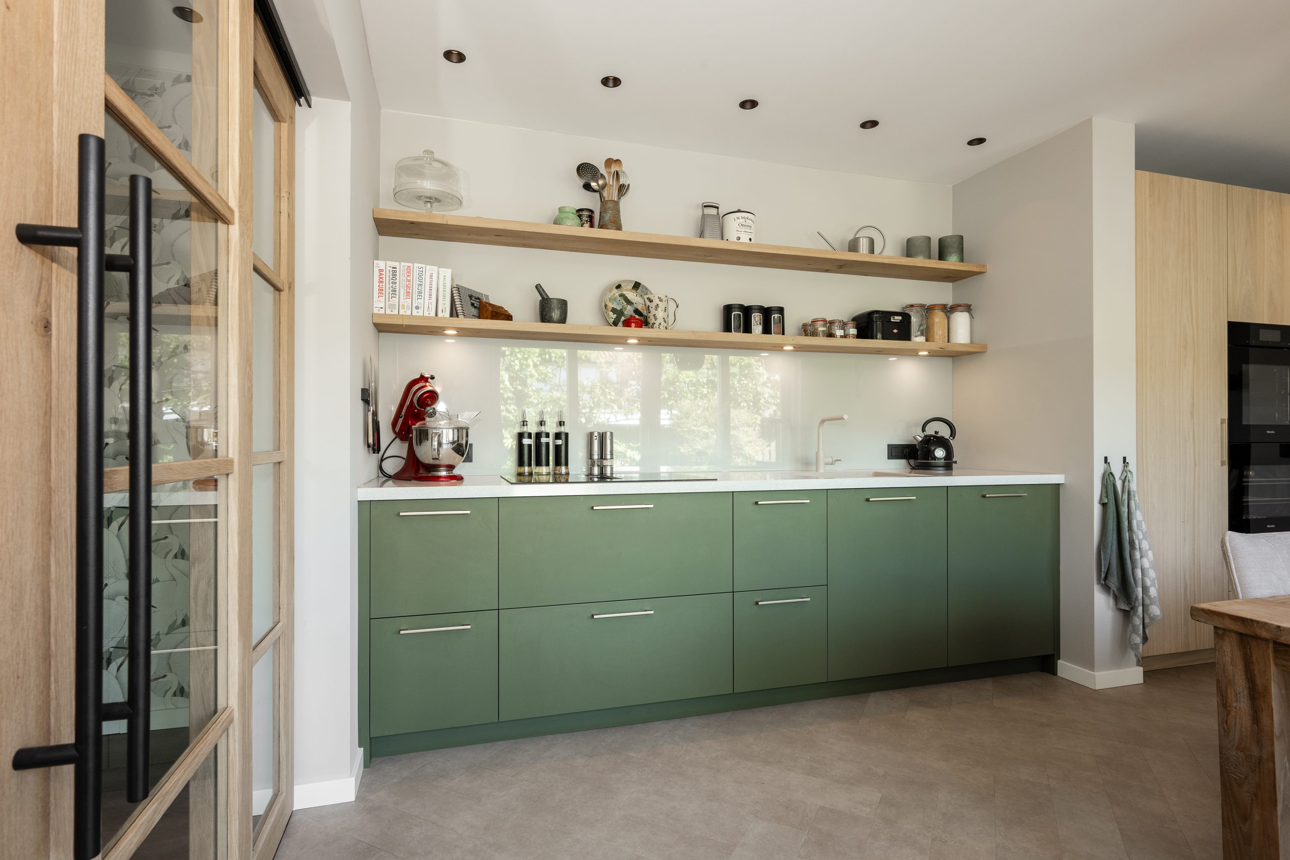 Keukenhoek met groene kasten, houten wandplanken en geïntegreerde oven in een hoge kastenwand.