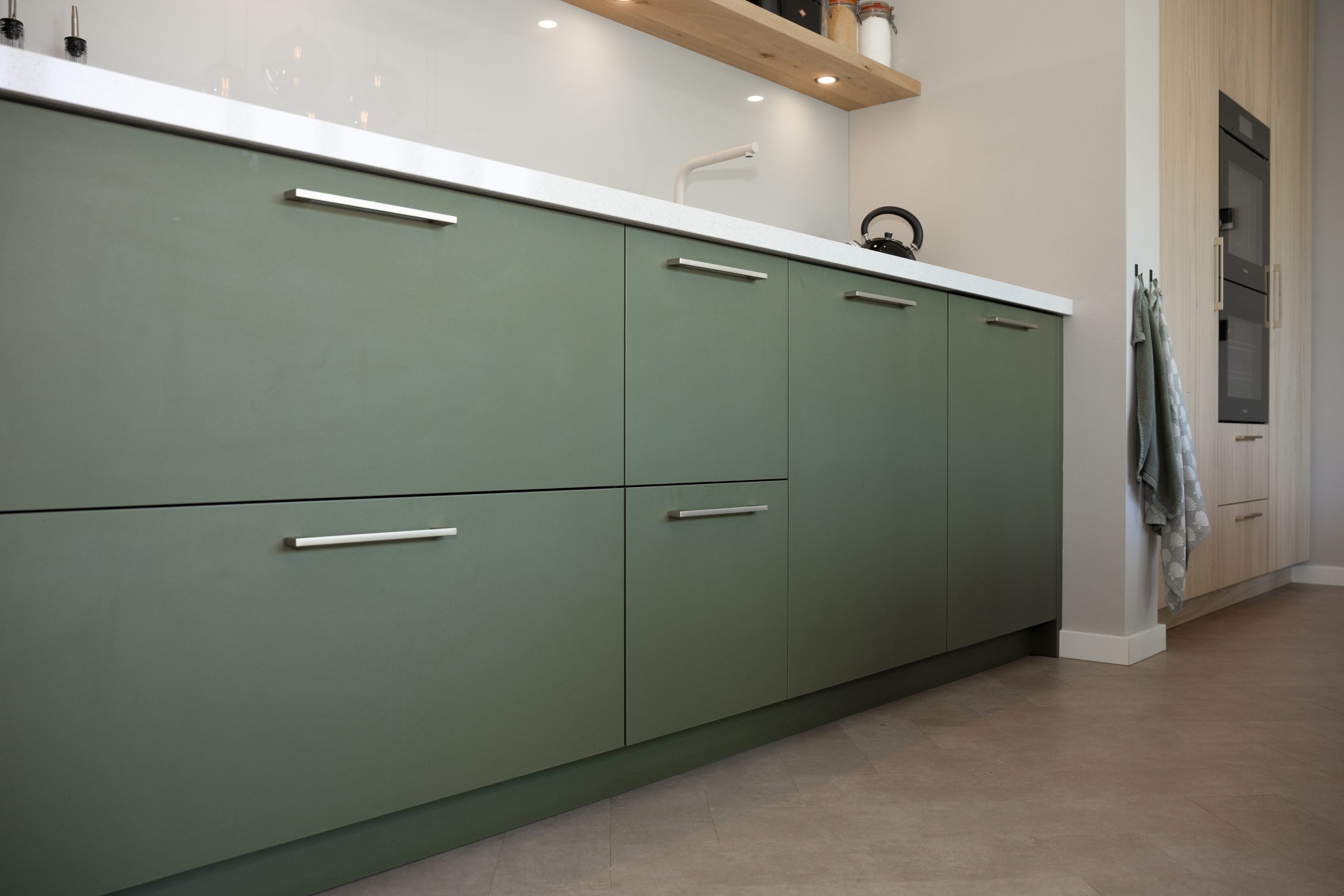 Zijaanzicht van een moderne keuken met groene kasten, wit aanrechtblad en een hoge houten kastenwand.
