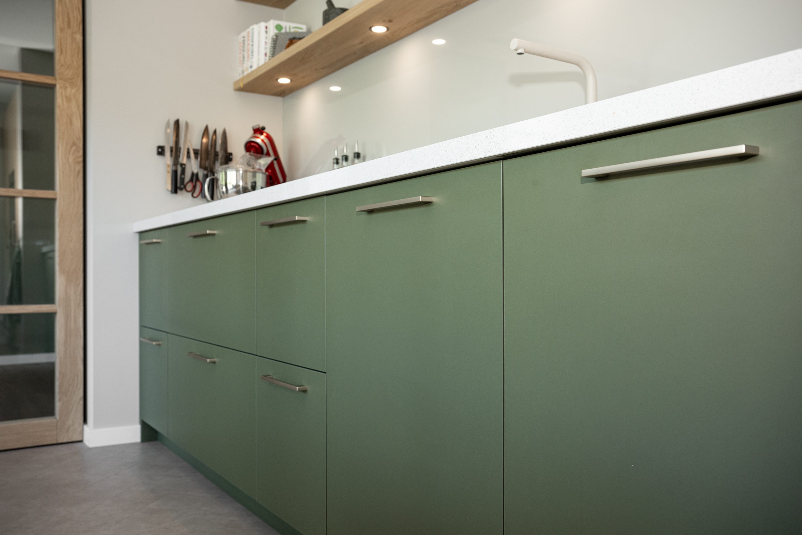 Schuin aanzicht op het keukenblok met groene fronten, wit aanrecht en een wand met messenblok en keukenmachine.