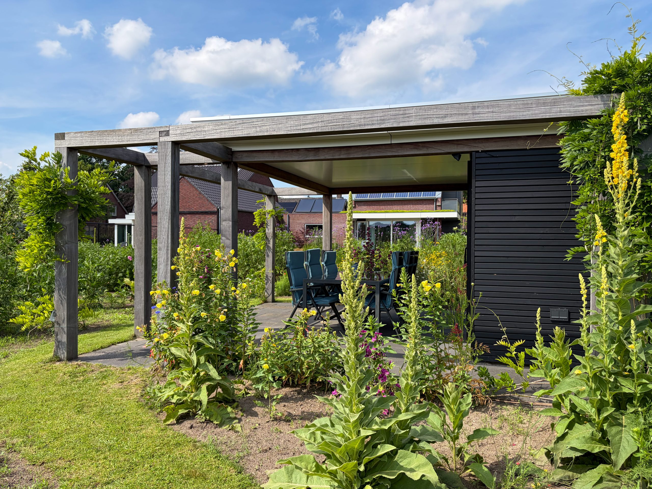 Zonnig overzicht van een moderne houten overkapping met weelderige gele bloemen en beplanting op de voorgrond.