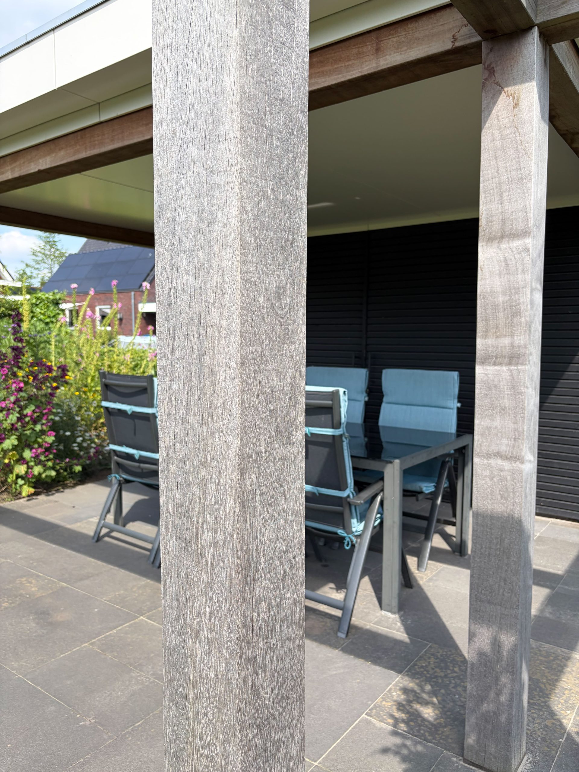 Scherpe opname van een houten staander van een overkapping met daardoorheen zicht op de tuinstoelen en tafel.