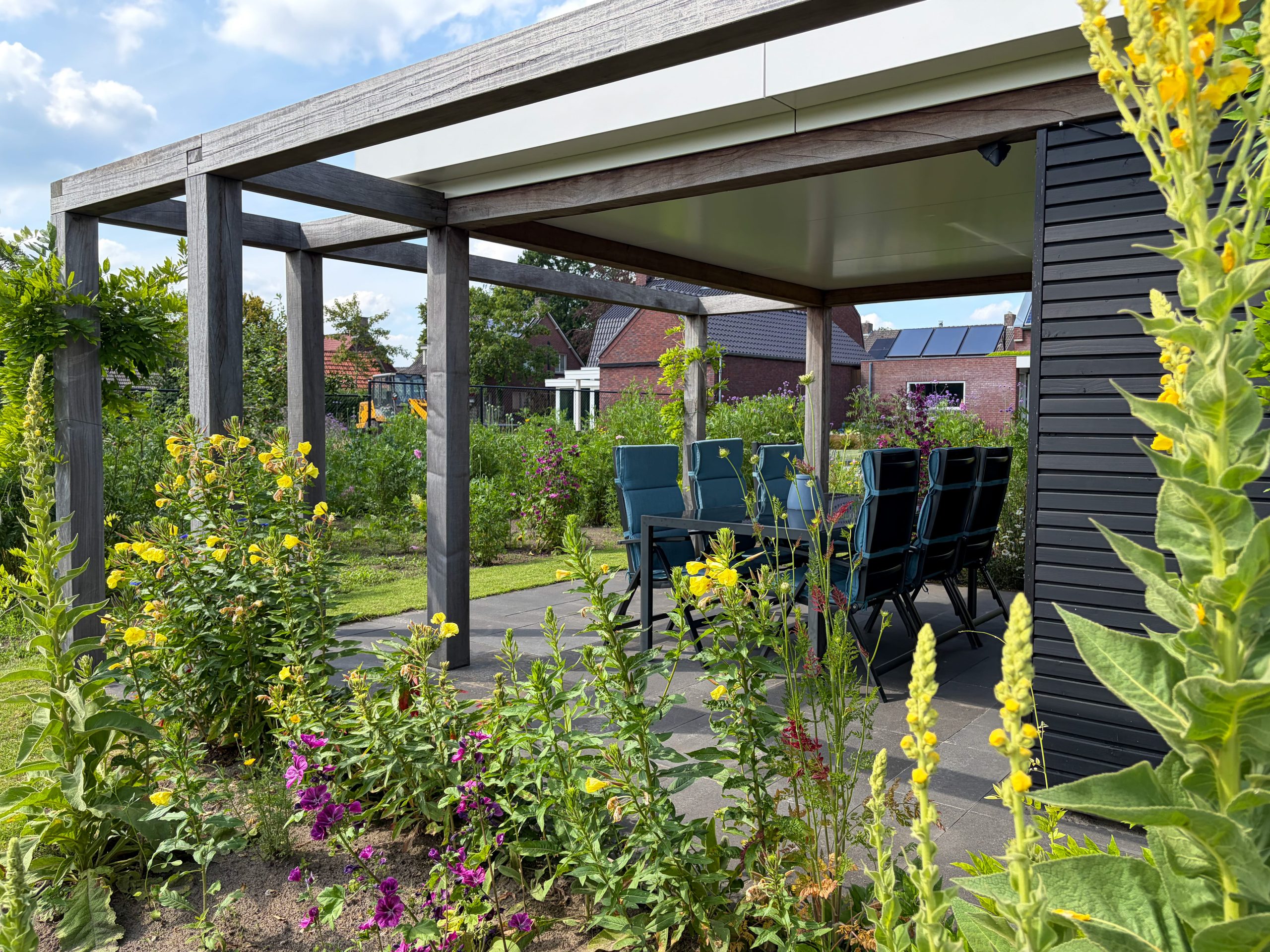 Luxe houten overkapping met een tuinset, omringd door hoge gele en paarse bloeiende planten in de tuin.