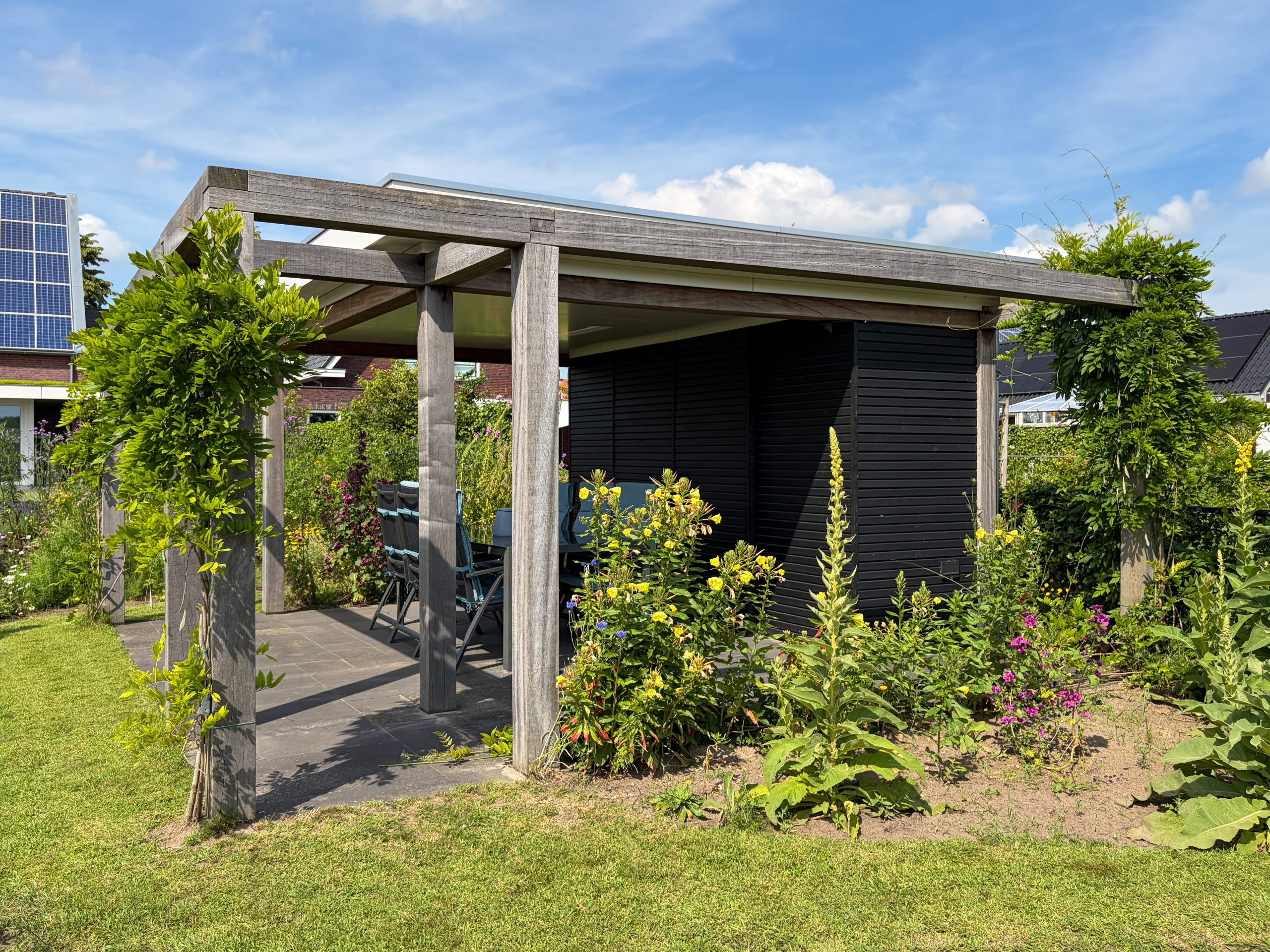 Moderne houten pergola en tuinhuis met gele en roze bloemen op de voorgrond tijdens een zonnige zomerdag.