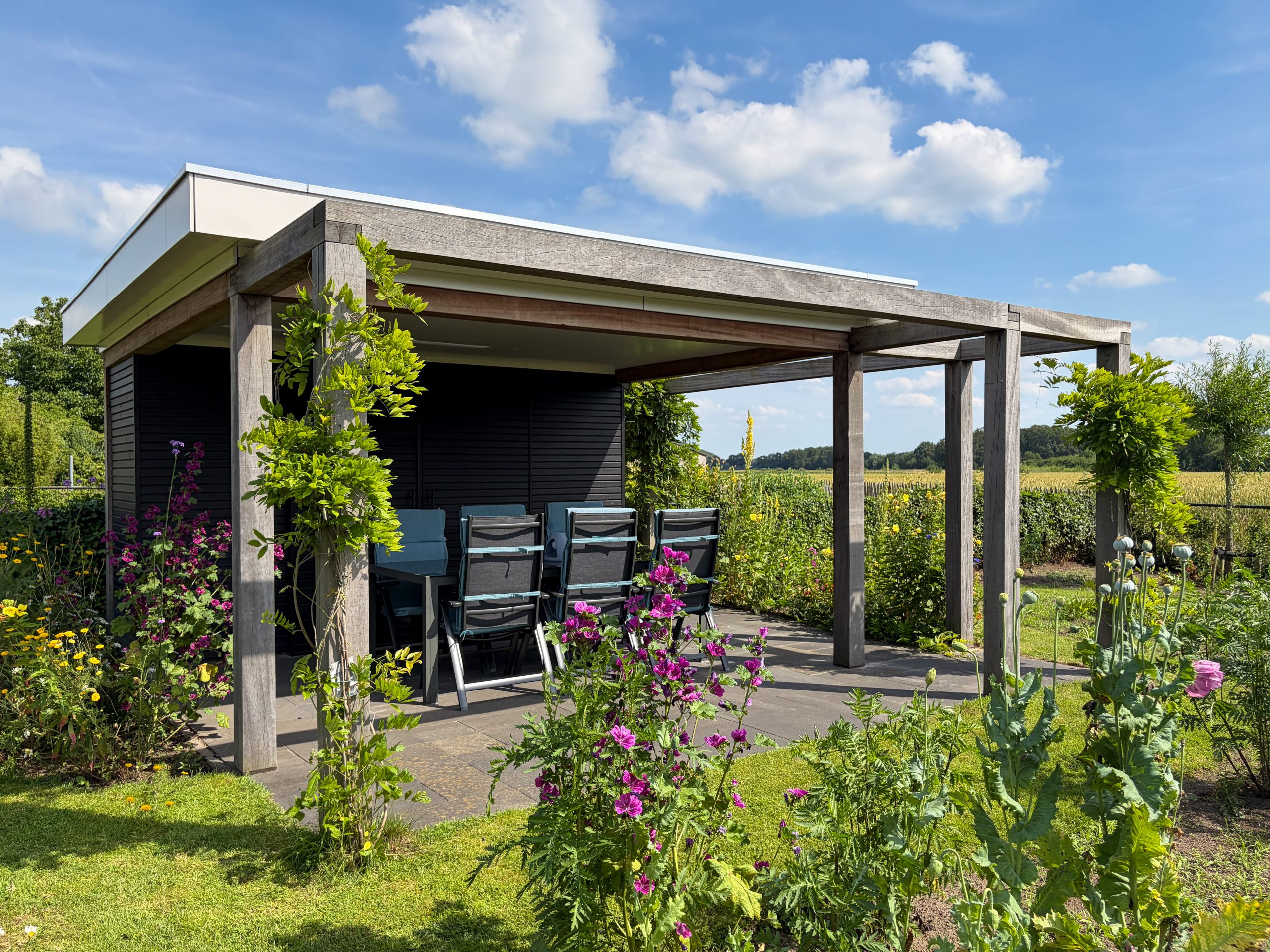 Luxe houten overkapping met lounge-set op een zonnige zomerdag, staand te midden van een bloeiende natuurlijke tuin.