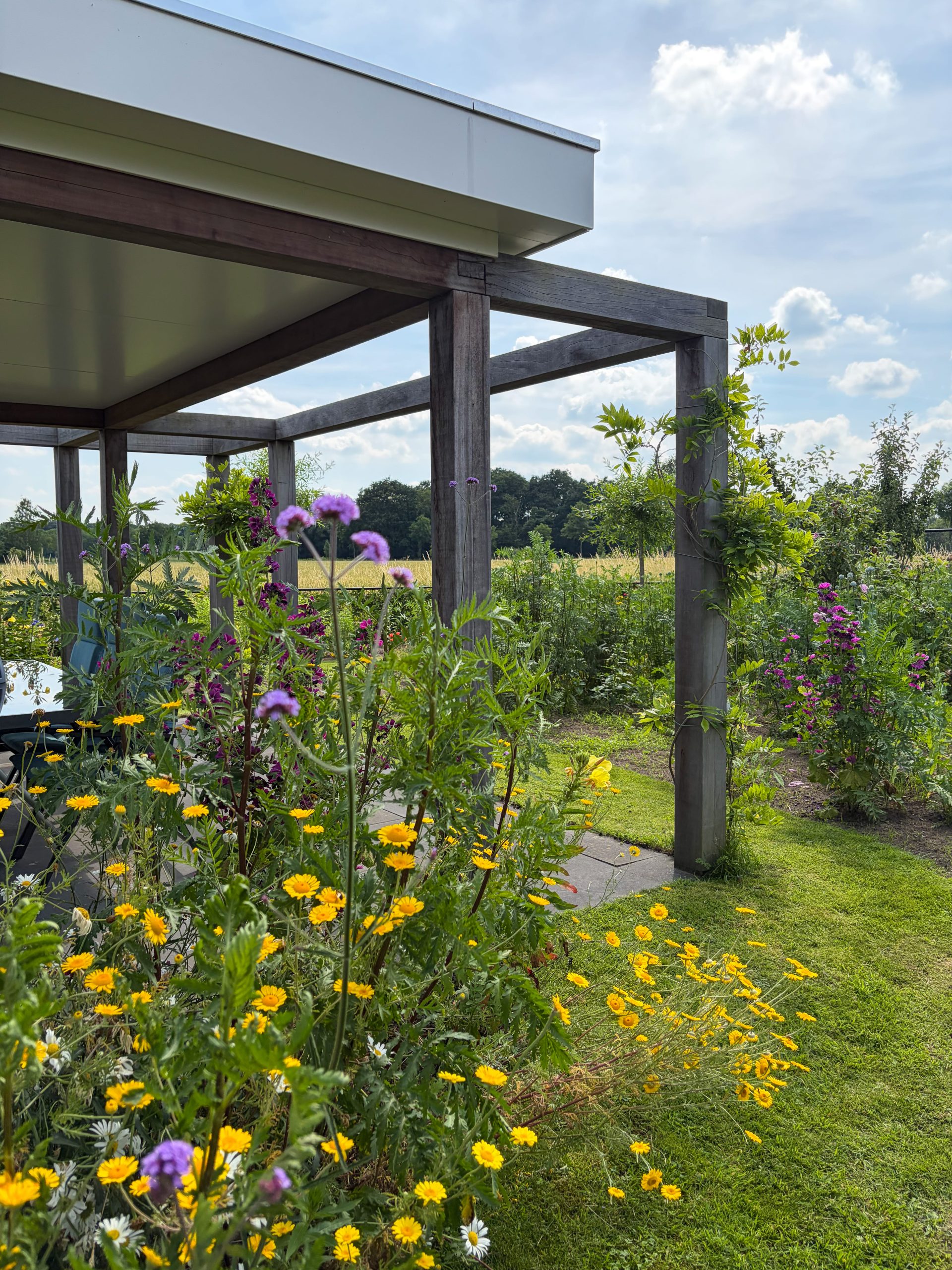 Zicht vanuit een houten overkapping op een weelderige tuin met gele en paarse bloemen en een landelijk landschap op de achtergrond.