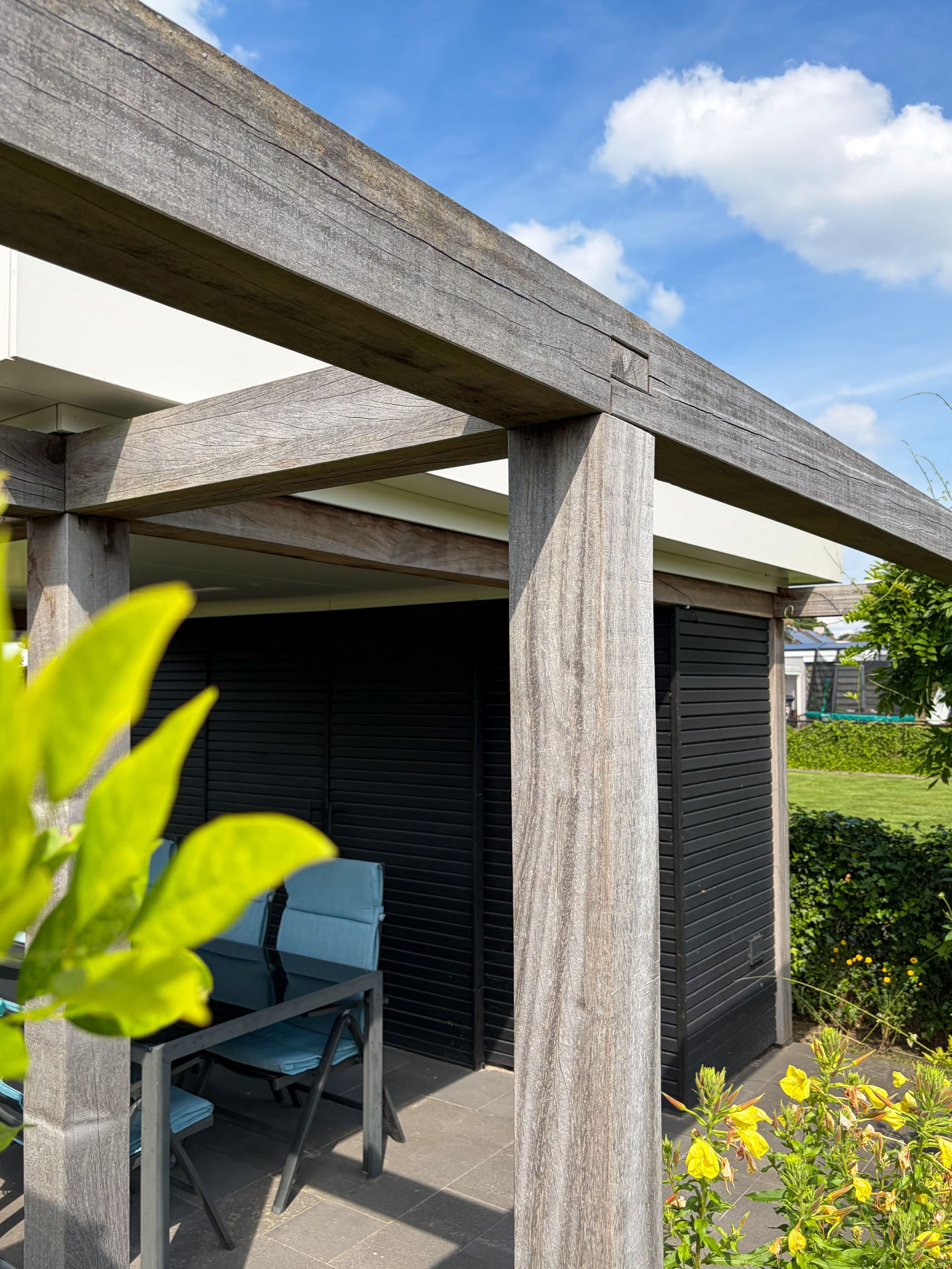 Detailopname van de houten balkenconstructie van een pergola met op de achtergrond een zonnige tuin.