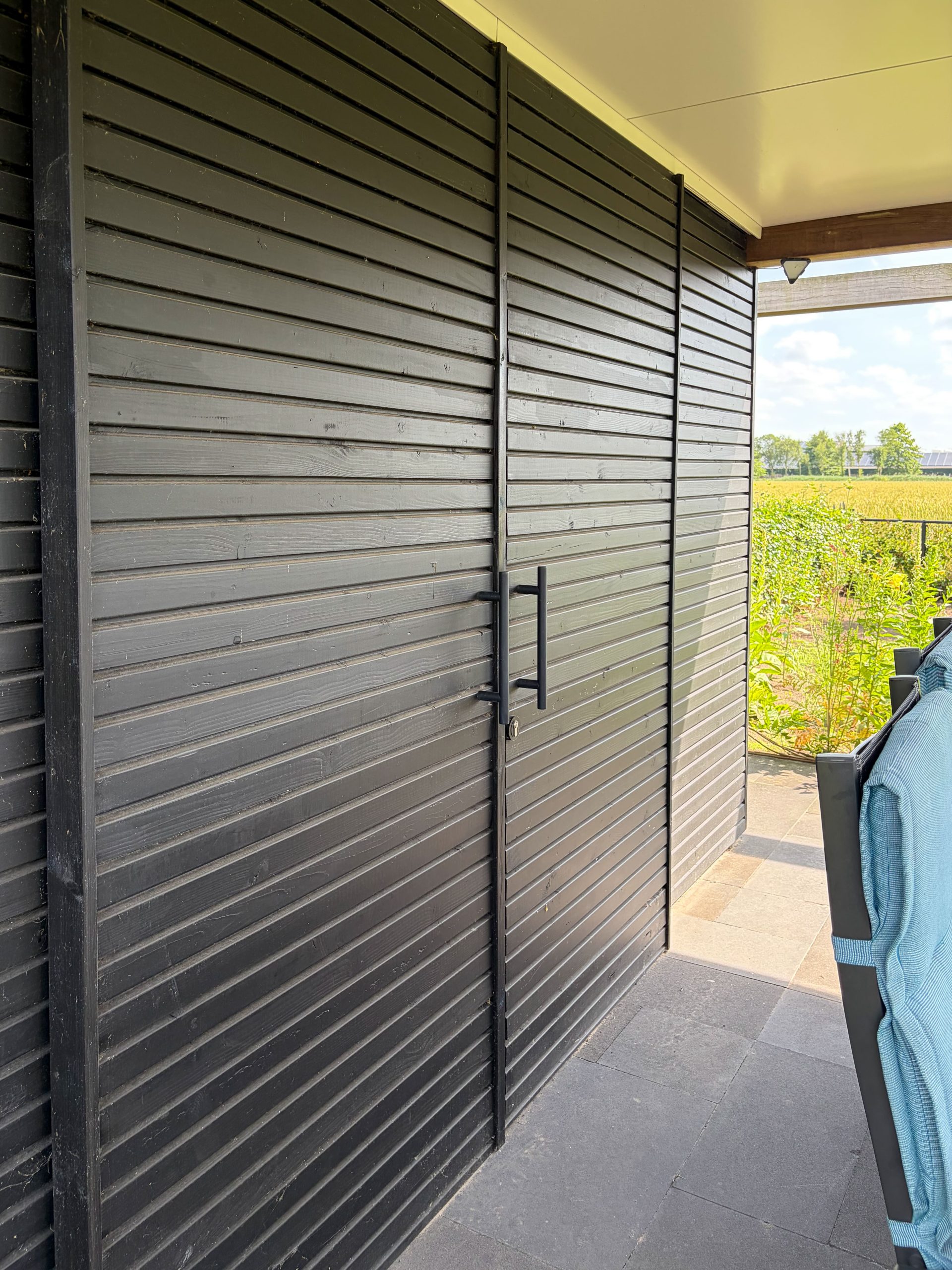 Detailopname van de zwarte houten gevelbekleding en schuifdeur van een moderne tuinhuis in een landelijke tuin.